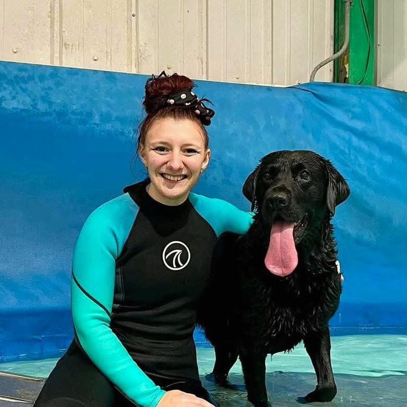 Chloe, hydrotherapist, in a wetsuit sitting in the pool with a black Labrador