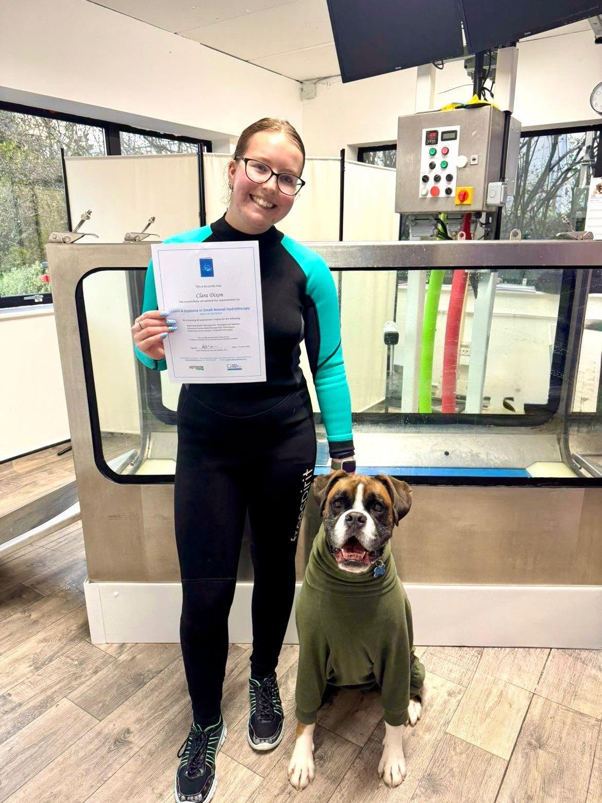 Clara holding her Level 4 Certificate in Canine Hydrotherapy beside the water treadmill, with Jasper the Boxer in a green jumper sitting next to her